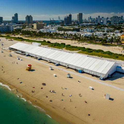 Aerial view of a big tent on the beach with people walking by and the city in the background.