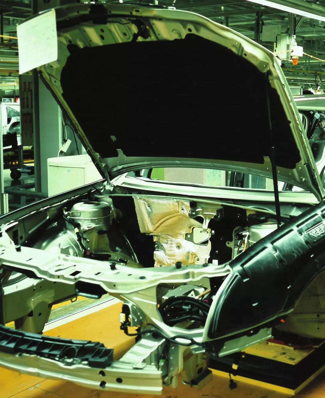 A brightly lit, close-up interior shot of an automotive assembly line, showing the metal frame, and open door and hood of a car body being constructed.