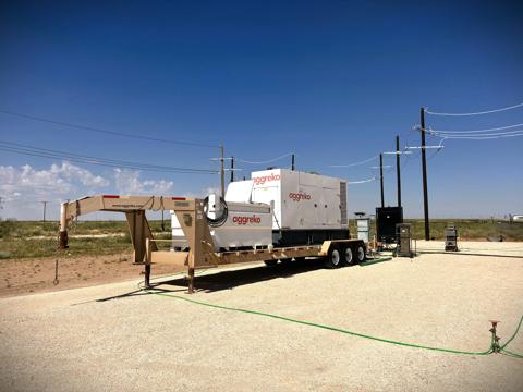 A white, containerized industrial Aggreko generator unit sits on a brown, multi-axle trailer in an open field.
