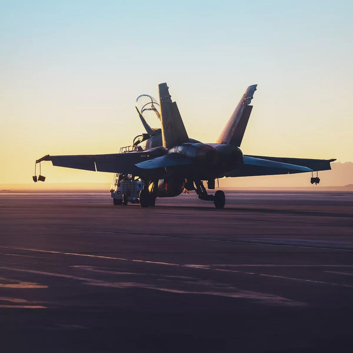 A low-angle exterior view of a military fighter jet, sitting on a runway during sunset with the canopy opened.
