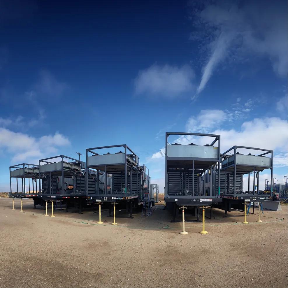 A panoramic view of a dusty, open plain at the base of a large, arid, rocky mountain range. On the right, several industrial container units are mounted high on trailers in the foreground.