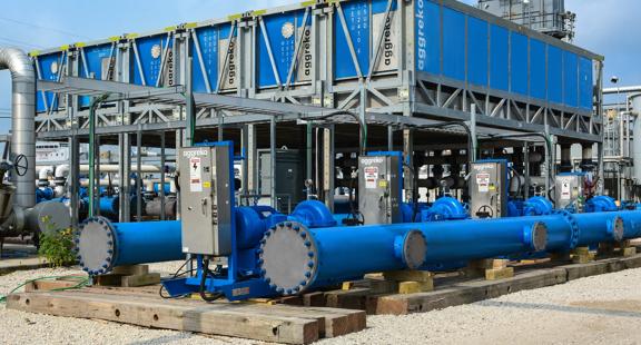 A ground-level shot of large, bright blue industrial pipes and pumps in front of stacked blue Aggreko containers.