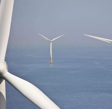 Three wind turbines stand in the ocean, with their tall structures rising above the water's surface in a clear blue sky.