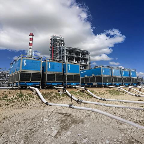 Several blue industrial units stacked two-high in an arid, outdoor setting with large white and black hoses connected to the bottom, and other industrial structures visible in the background under a blue, cloudy sky