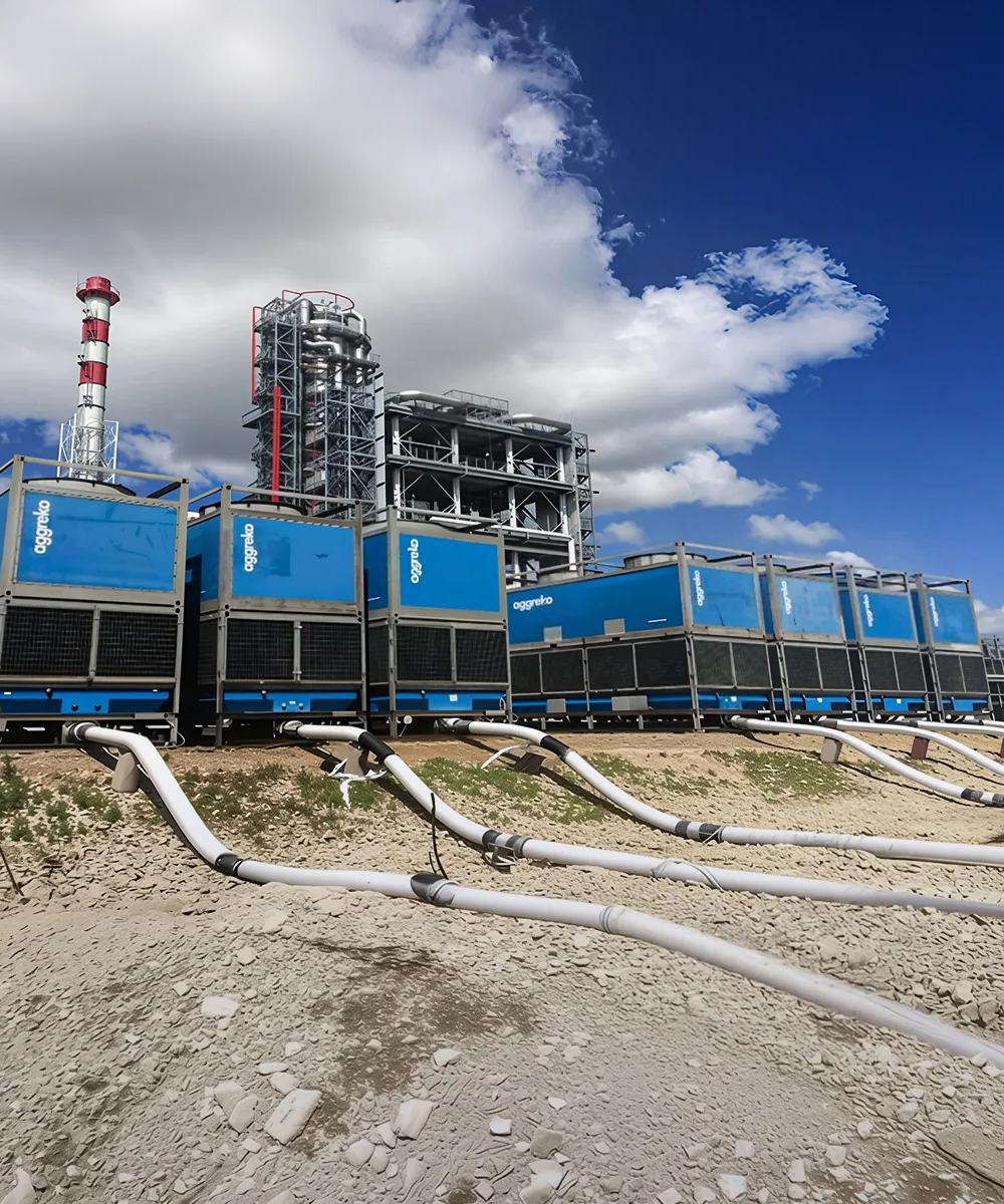 Several blue industrial units stacked two-high in an arid, outdoor setting with large white and black hoses connected to the bottom, and other industrial structures visible in the background under a blue, cloudy sky