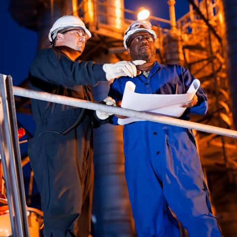 View of 2 workers looking at a blueprint at a refinery setting.