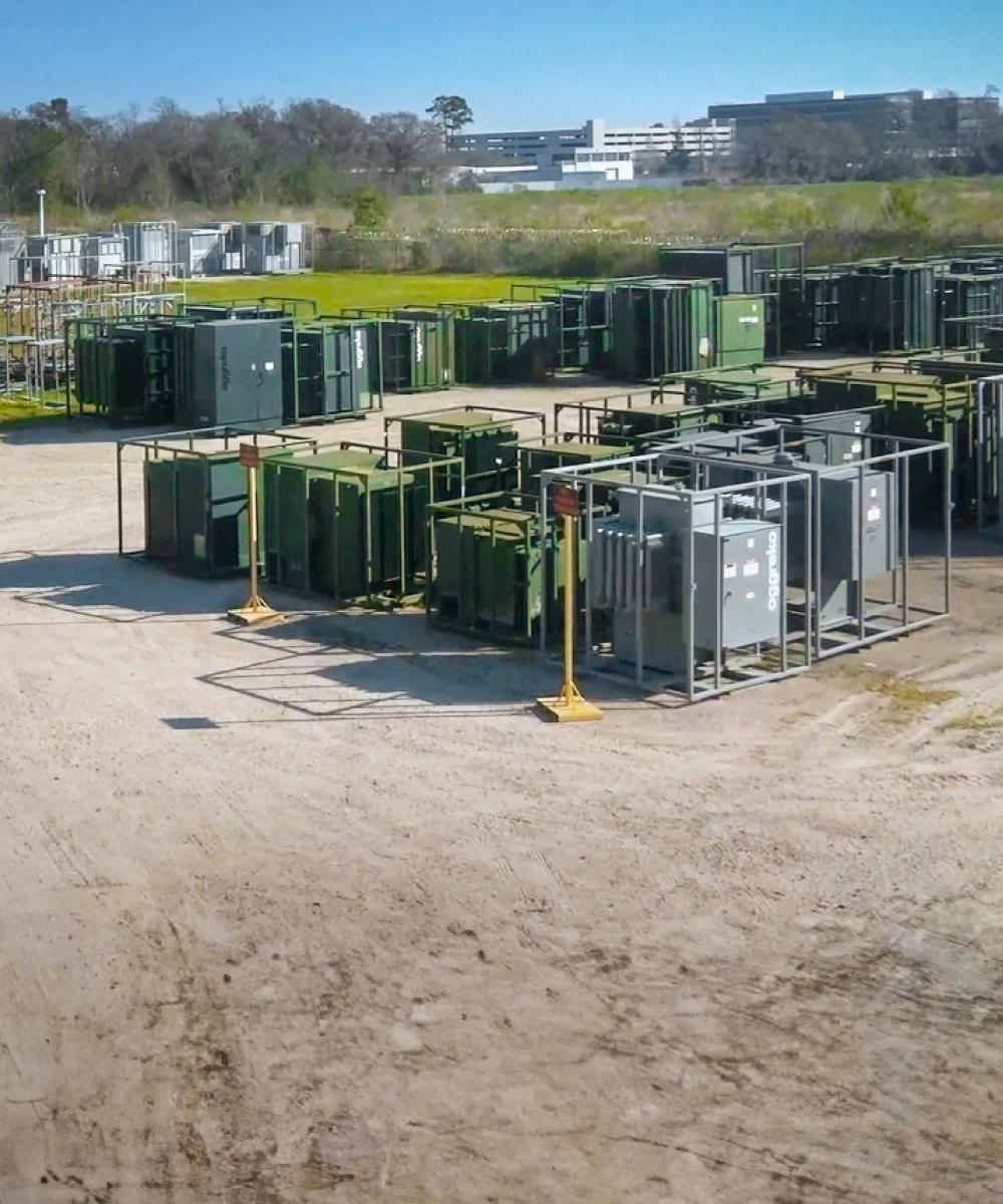 View of multiple gray and green transformers in an industrial lot.