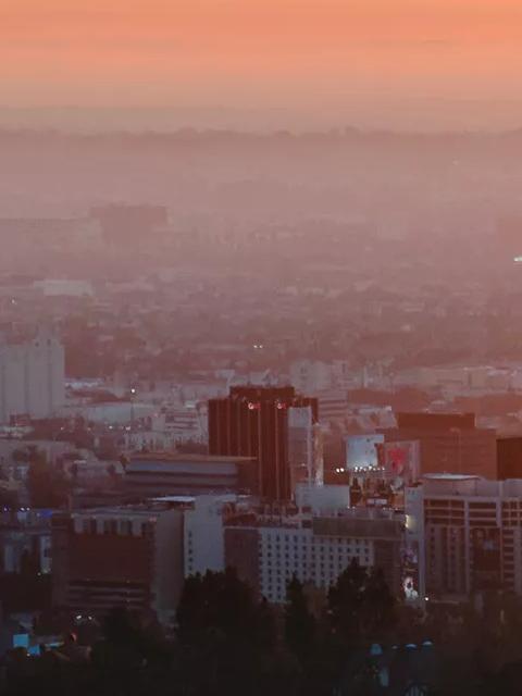 A wide, elevated view of a large city skyline, partially obscured by low-hanging fog, dramatically lit by a warm, orange and yellow sunrise.