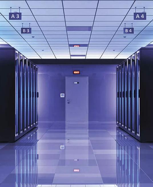 A long, symmetrical perspective view down an aisle in a massive server room, with towering rows of racks illuminated by a cool blue light from vertical LED strips, and the floor reflecting the light.