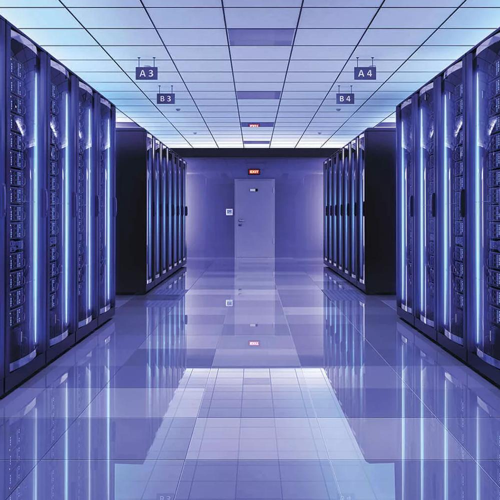 A long, symmetrical perspective view down an aisle in a massive server room, with towering rows of racks illuminated by a cool blue light from vertical LED strips, and the floor reflecting the light.