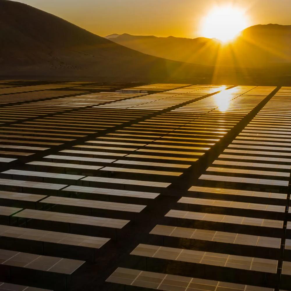Wide shot of a row of solar panels with mountains and the Sun in the background, during a yellow sunset.