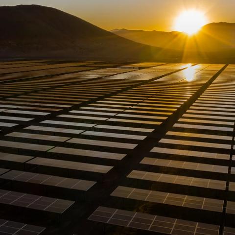 Wide shot of a row of solar panels with mountains and the Sun in the background, during a yellow sunset.