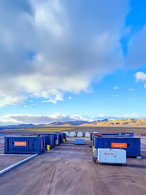 Several industrial Aggreko units on a dirt lot, with a rocky hillside in the background.