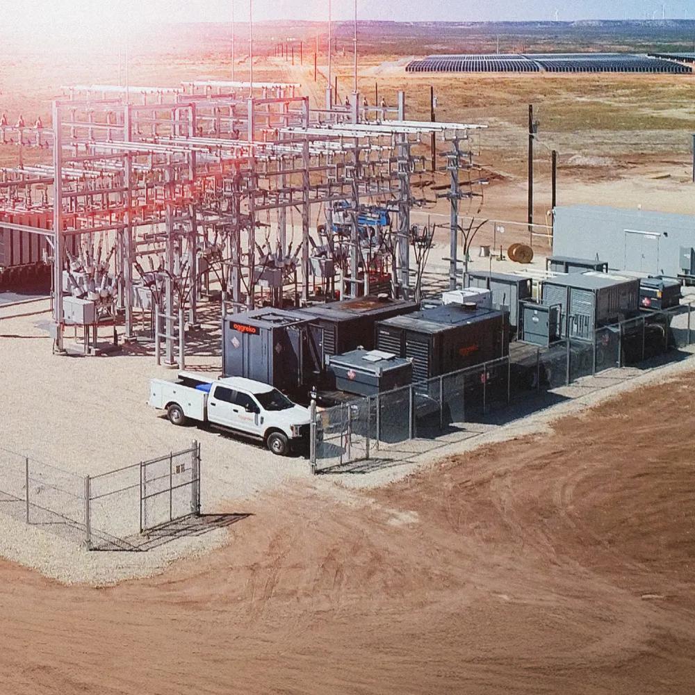 An aerial view of a large utility-scale solar project with extensive rows of solar panels covering the left half of the image in a dry, open field. On the right, a complex electrical substation with large metal structures and transformers, with a white truck parked inside the fenced perimeter.