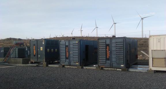 A row of three black Aggreko generators in gravel yard, a hill behind them, and multiple wind turbines behind the hill