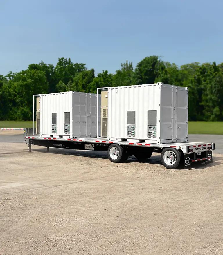 A truck with two large white generators is parked in an empty, gravelly area surrounded by green trees.