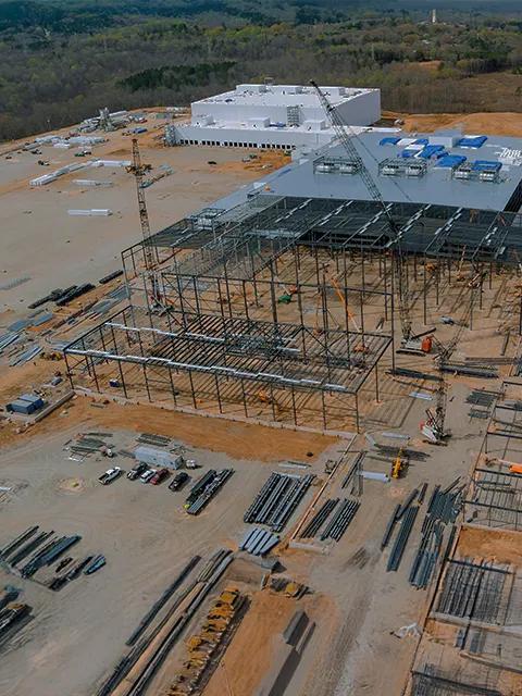 An aerial view of a massive data center construction site. The foreground shows the steel skeleton of a building under construction, with half of it being partially completed. A large, white completed building stands in the background. Cranes and construction materials are scattered across the surrounding dirt site.