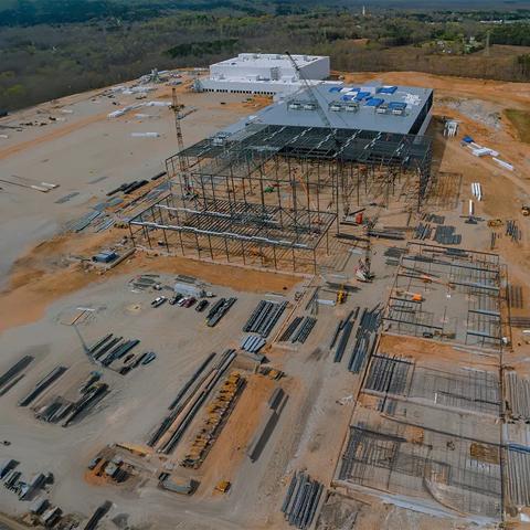Une vue aérienne d’un chantier de construction de centre de données massif. Au premier plan, on voit la structure d'acier d'un bâtiment en construction, dont la moitié est partiellement complétée. Un grand bâtiment blanc complété se dresse à l'arrière-plan. Des grues et des matériaux de construction sont dispersés sur le site en terre tout autour.