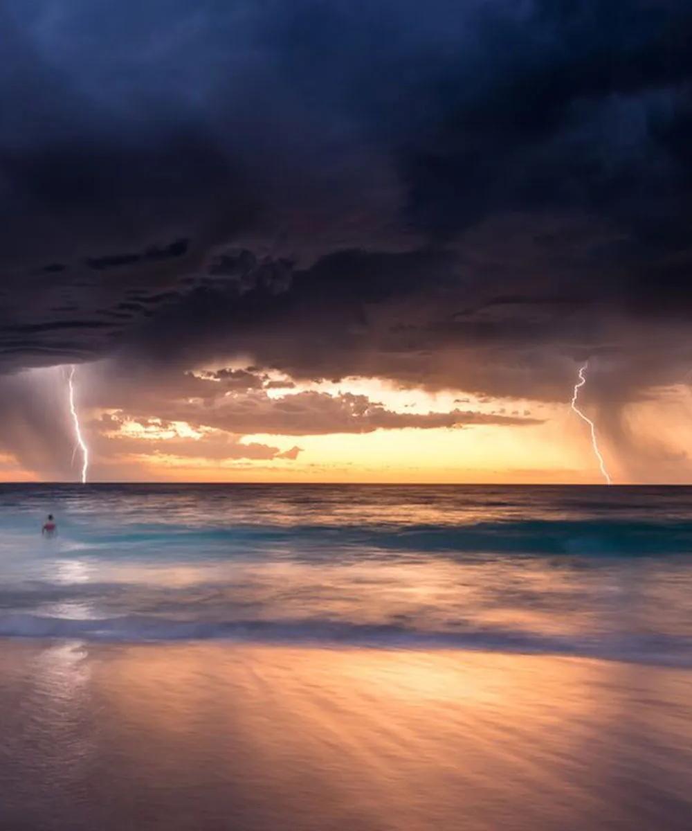 A dramatic image of a dark, stormy sky over the ocean with a bolt of lightning striking the water near the horizon, set against a bright yellow and orange glow at sunset.