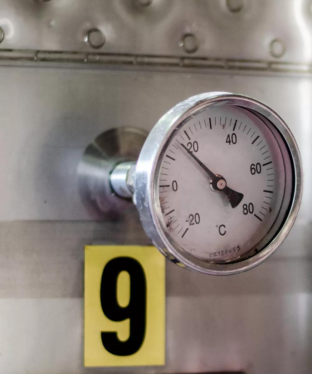 A close-up of a circular industrial thermometer gauge mounted on the side of a large, shiny stainless steel tank. The gauge needle points near 20°C, and a large yellow label with the number "9" is affixed below it.