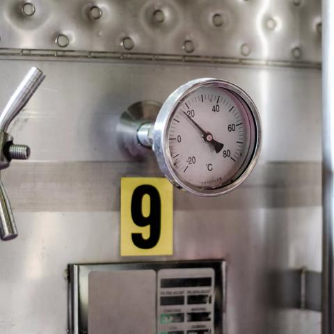 A close-up of a circular industrial thermometer gauge mounted on the side of a large, shiny stainless steel tank. The gauge needle points near 20°C, and a large yellow label with the number "9" is affixed below it.