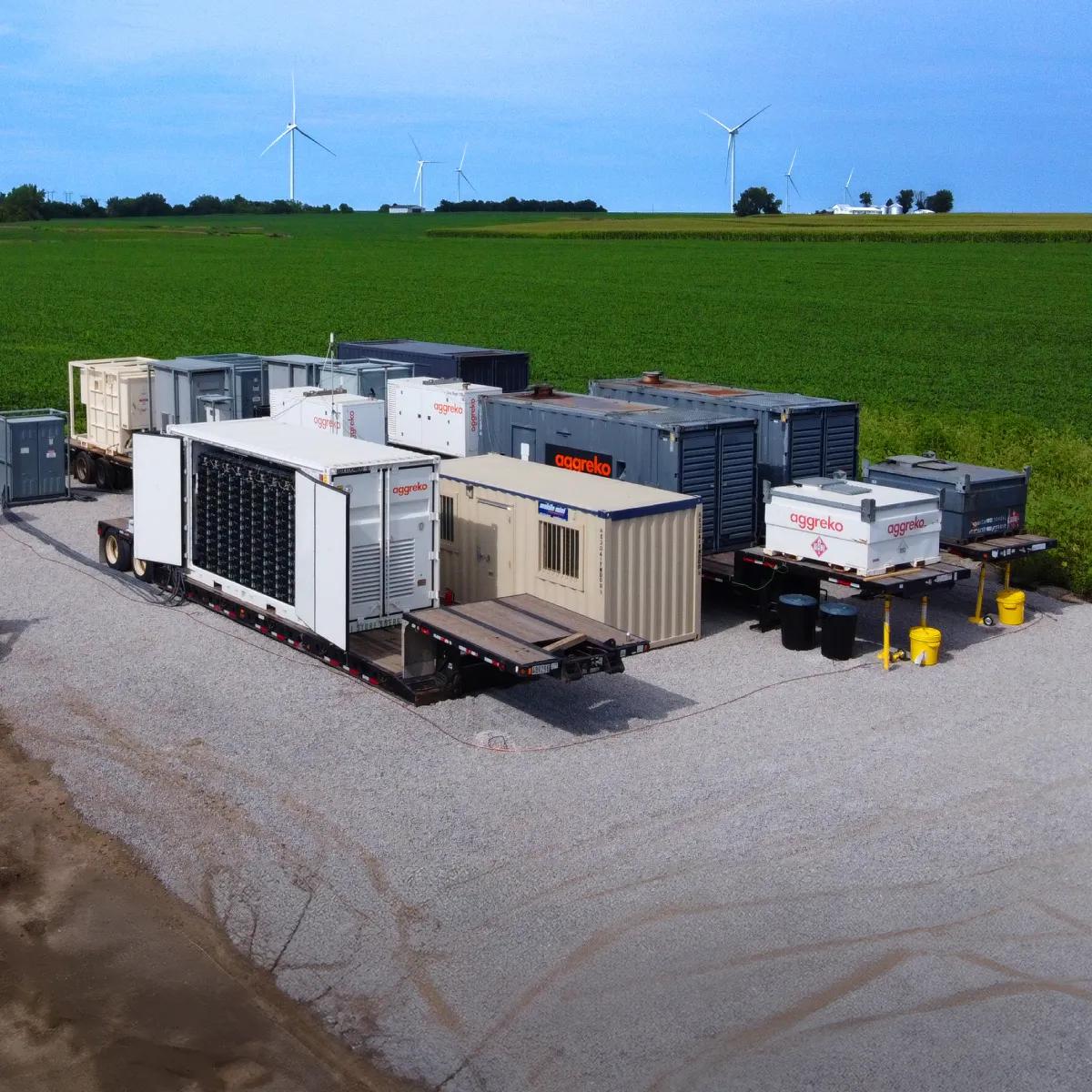 A dirt road leads through a green field, with gas generators and equipment on the side, near wind turbines in the distance.