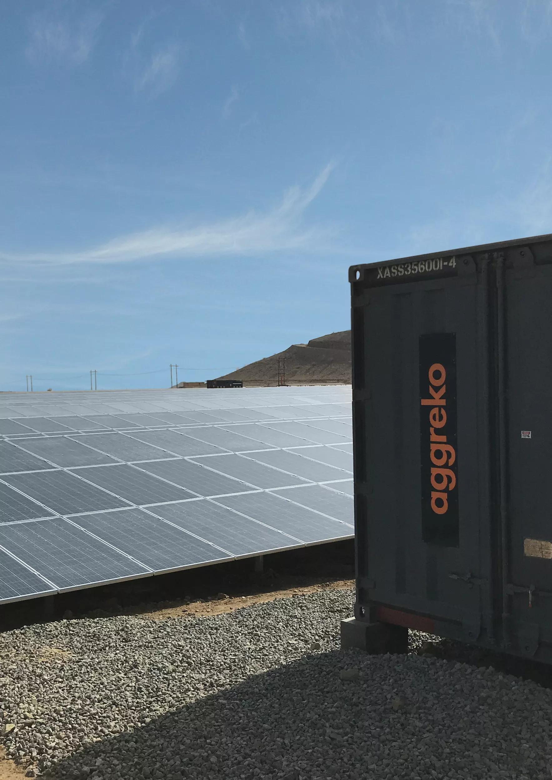 A large field of solar panels and a dark Aggreko unit on a gravelly surface.
