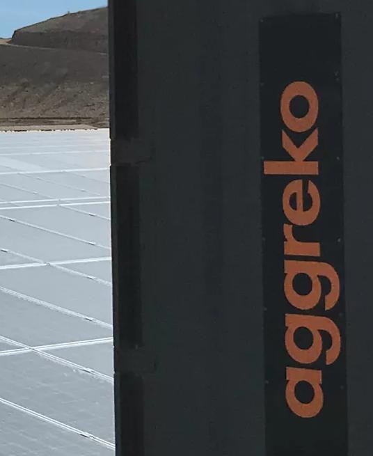 A large field of solar panels and a dark Aggreko unit on a gravelly surface.