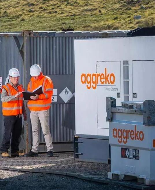 A row of white and gray Aggreko modular units and a small building are situated at the base of a large, barren, brown hill.