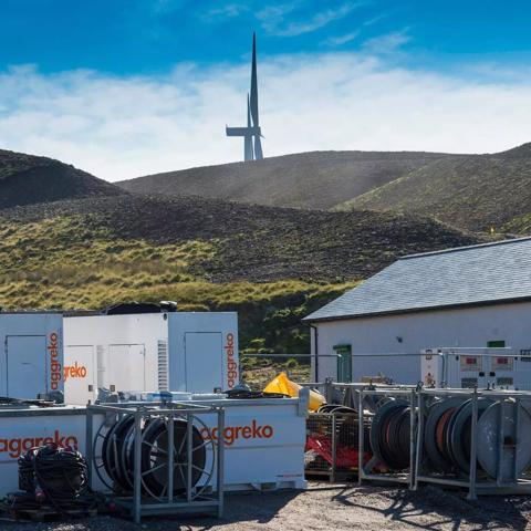 A row of white and gray Aggreko modular units and a small building are situated at the base of a large, barren, brown hill.