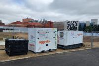 A white containerized Aggreko generator unit sits on a paved surface near industrial buildings.