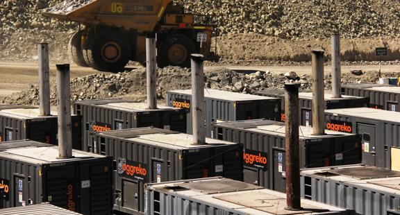 A large yellow mining truck driving past a collection of black industrial Aggreko units with exhaust pipes