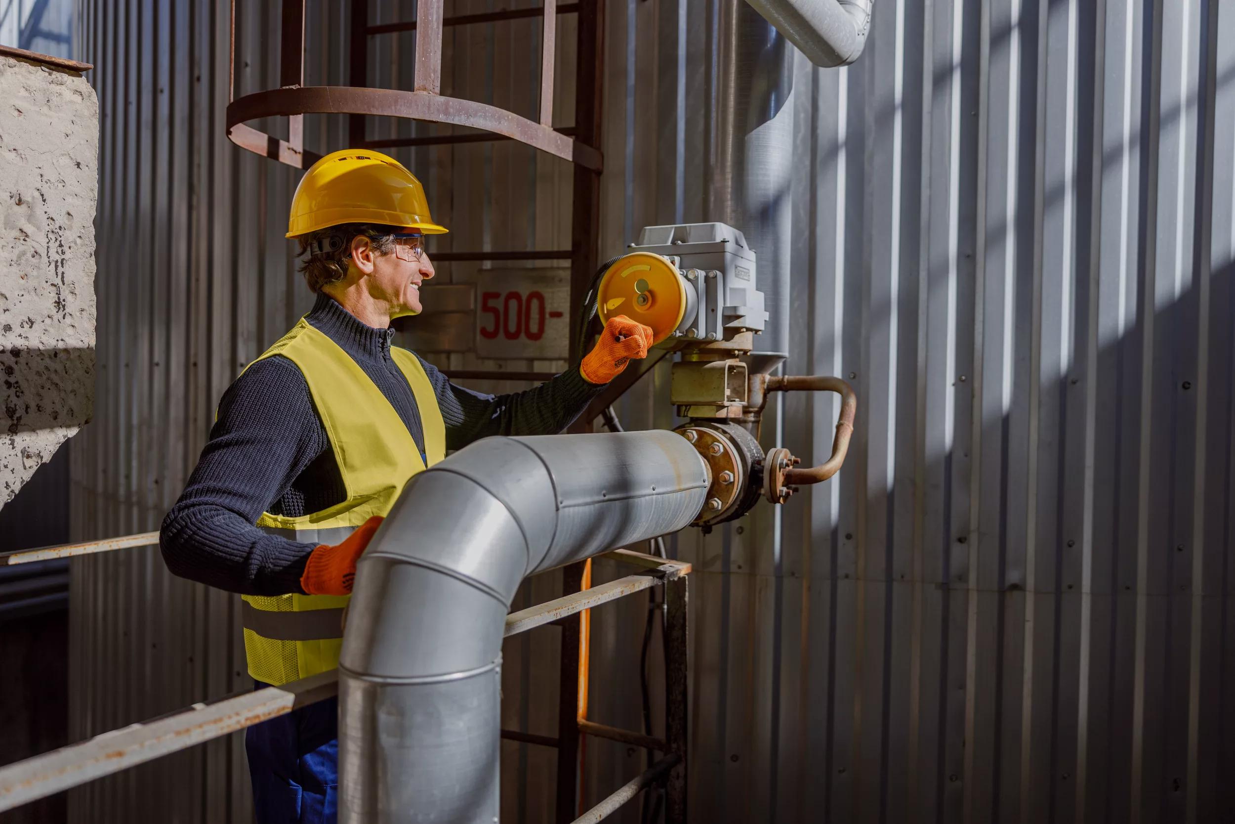 Cheerful matured man factory worker wearing work vest and gloves while standing near metal pipe and smiling