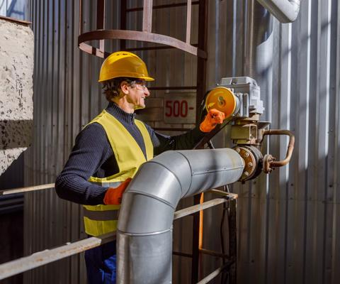 Cheerful matured man factory worker wearing work vest and gloves while standing near metal pipe and smiling