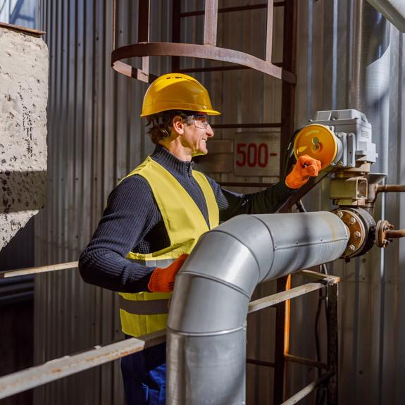 Cheerful matured man factory worker wearing work vest and gloves while standing near metal pipe and smiling