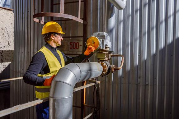 Cheerful matured man factory worker wearing work vest and gloves while standing near metal pipe and smiling