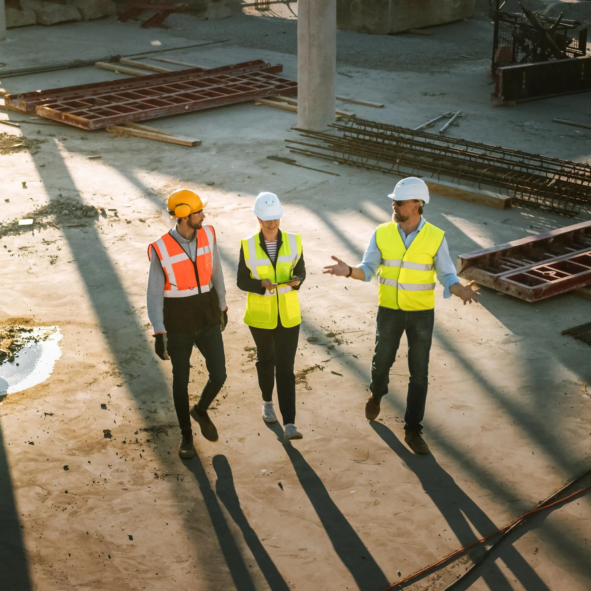 Three construction workers in safety gear walk through a building site filled with concrete columns and machinery.