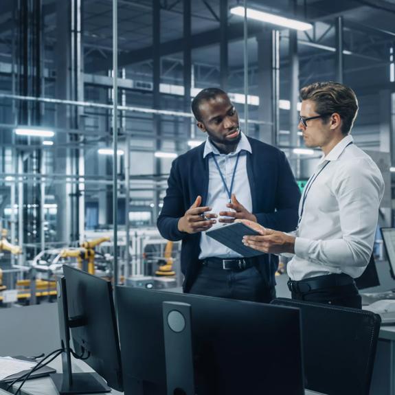 Two Diverse Automotive Industrial Engineers Talking About Vehicle Production while Standing in Office at a Car Assembly Plant. Industrial Specialists Discuss Work Projects on Tablet Computer.