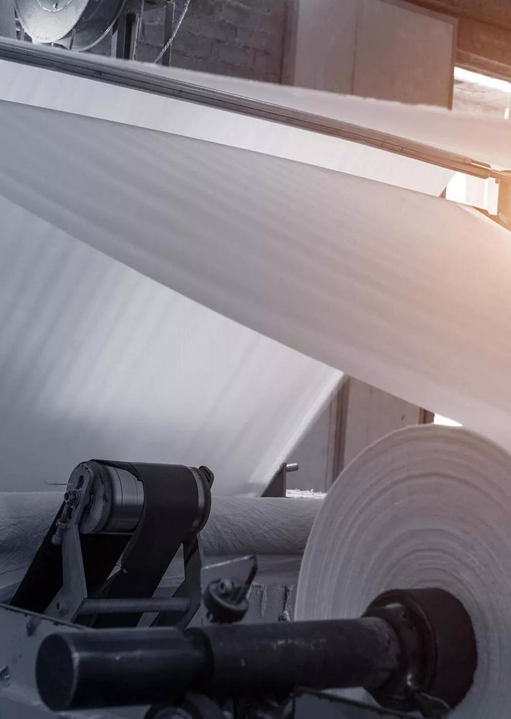 A wide, close-up shot showing a large, continuous sheet of white material being fed through industrial rollers in a factory.