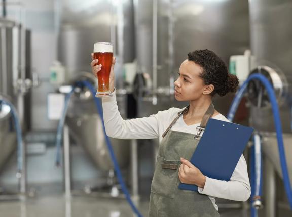 Waist up portrait of young African-American woman holding beer glass while inspecting quality of production at brewing factory, copy space