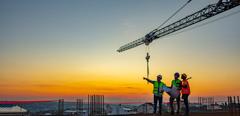 Three Multi-Ethnic construction workers in uniform standing at construction site with crane in background, discussing building plans while holding blueprint at sunset under the tower crane.