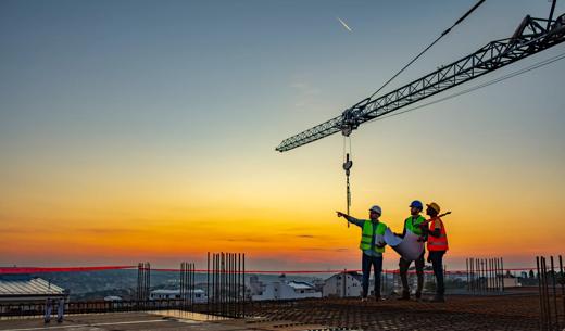 Three Multi-Ethnic construction workers in uniform standing at construction site with crane in background, discussing building plans while holding blueprint at sunset under the tower crane.