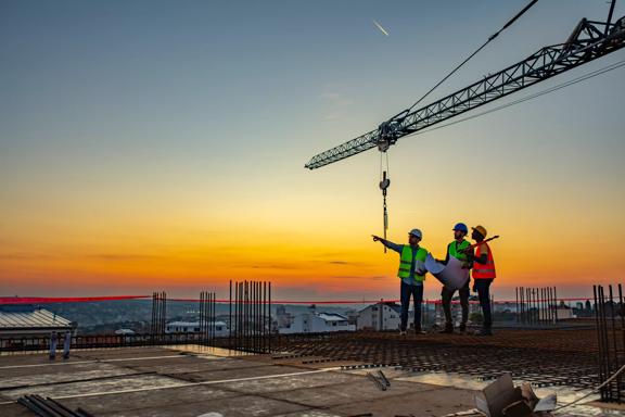 Three Multi-Ethnic construction workers in uniform standing at construction site with crane in background, discussing building plans while holding blueprint at sunset under the tower crane.