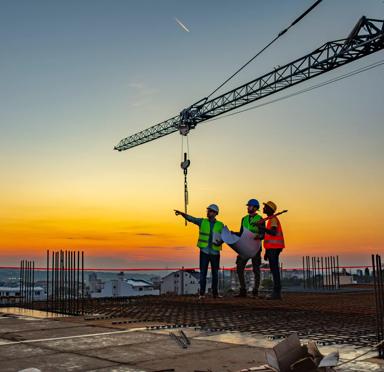 Three construction workers in hard hats and safety vests stand on a building site at sunset, discussing plans with a crane in the background.