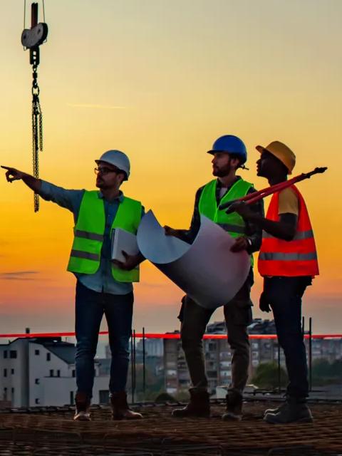 Three construction workers in hard hats and safety vests stand on a building site at sunset, discussing plans with a crane in the background.