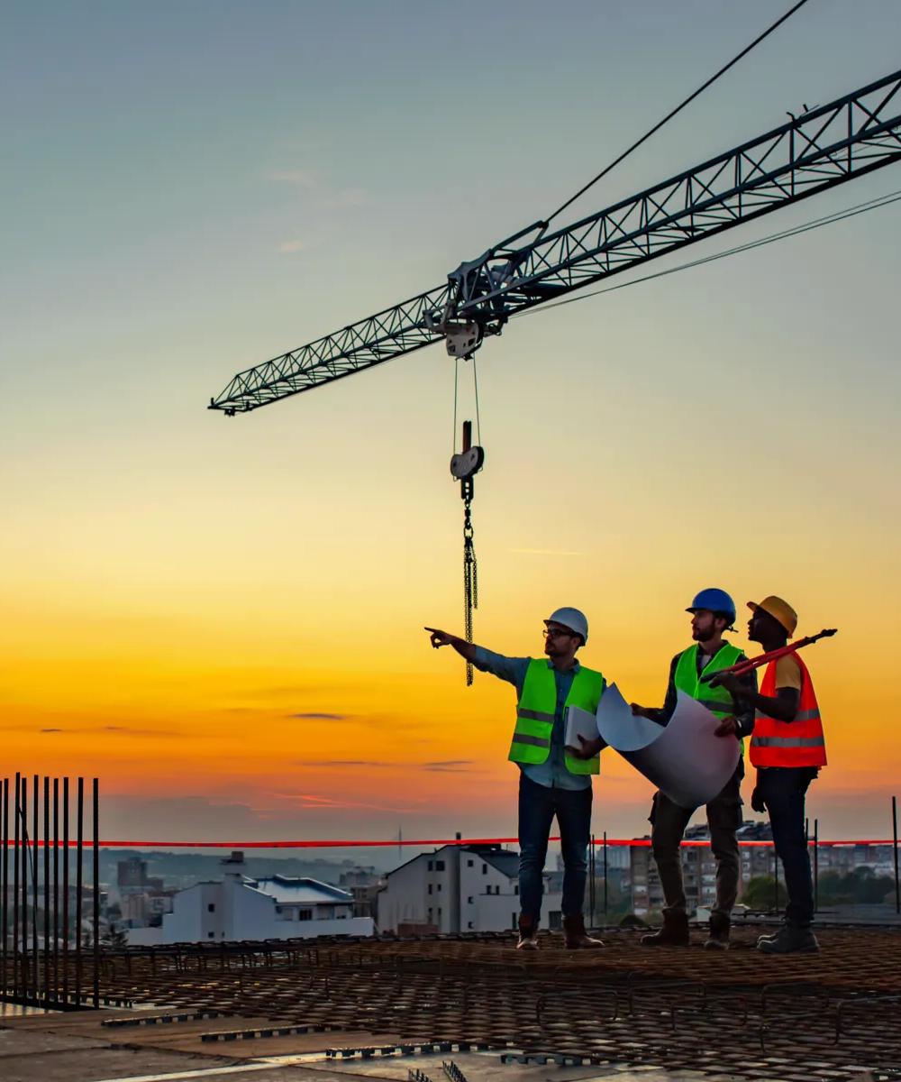 Trois travailleurs de la construction portant des casques et des gilets de sécurité se tiennent sur un chantier au coucher du soleil, discutant des plans avec une grue en arrière-plan.