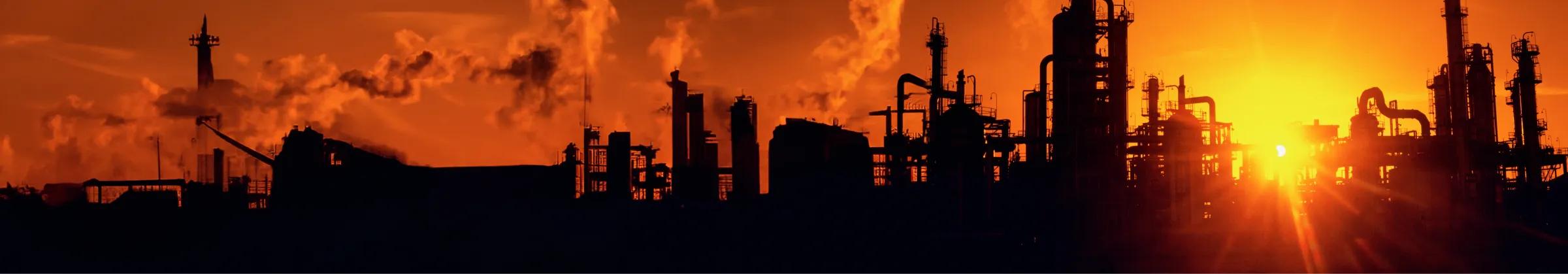 Silhouette of industrial structures against a vibrant orange sunset, with smoke rising from the facilities.