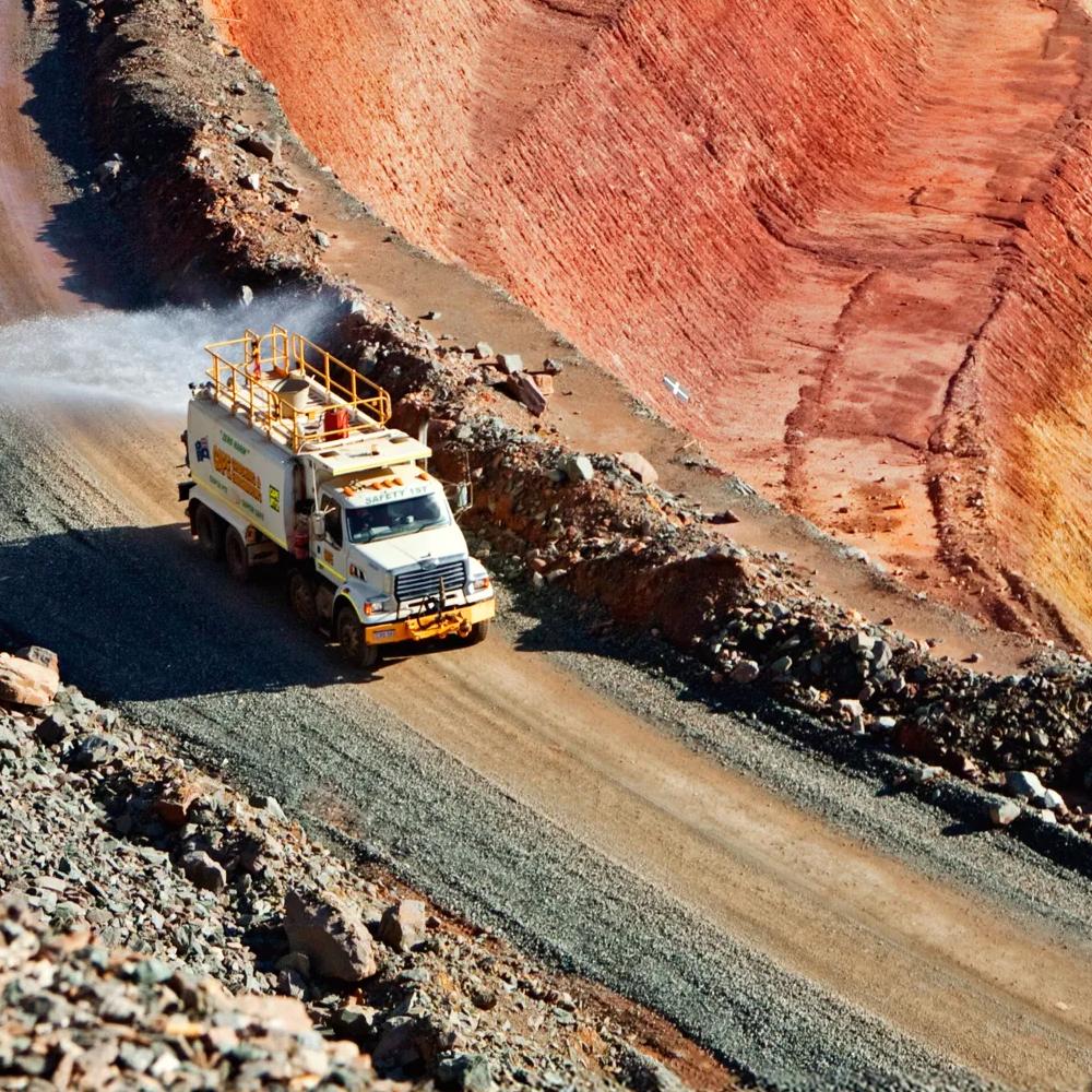 A haul truck driving on a curved, dusty road in an open-pit mine with deep red and brown earth.