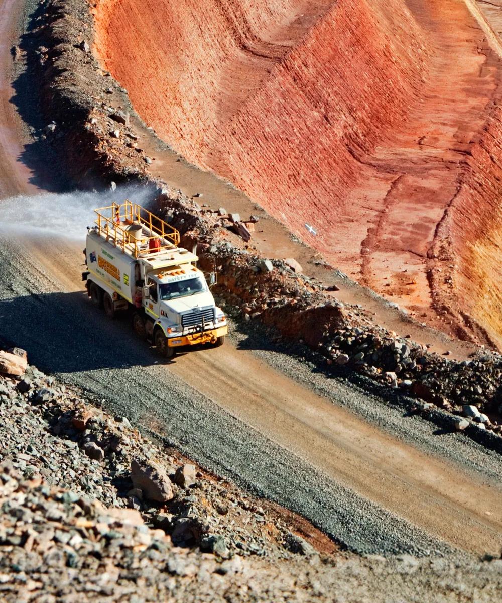 A haul truck driving on a curved, dusty road in an open-pit mine with deep red and brown earth.