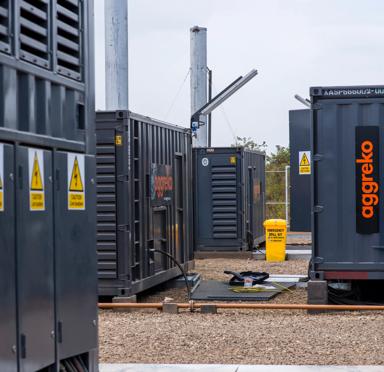 A black Aggreko generator with the orange logo in a gravel yard and equipment with caution signs